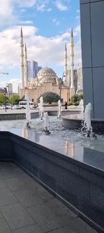 The media file captures a frontal view of the Akhmad Kadyrov Mosque in Grozny, Russia. The mosque, a prominent landmark, is characterized by its large central dome, multiple minarets, and ornate architecture. In the foreground, a modern fountain with several water jets adds a dynamic element to the scene. The water sprays upwards, creating a shimmering effect.

The setting appears to be an urban plaza or park area in front of the mosque. The time of day is daytime, indicated by the bright sunlight and the clear blue sky with scattered clouds. The weather seems pleasant, with no signs of rain or harsh conditions.

Several people are visible in the mid-ground, walking around the mosque and its surroundings. They are casually dressed, suggesting a normal day for visitors or residents. The presence of vehicles on a street behind the fountain and mosque, along with modern buildings in the background, places the scene firmly in a contemporary urban environment.

Notable details include the contrast between the traditional Islamic architecture of the mosque and the modern elements of the fountain and surrounding buildings. The lighting suggests it could be late morning or afternoon. The overall mood conveyed is one of peace and architectural grandeur, with the splashing water adding a sense of serenity. There is no visible text in the media file.
