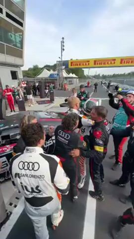 This video captures a celebratory scene at what appears to be a racetrack's pit lane, identified by the user as Monza, Italy. A group of approximately 15-20 individuals, primarily men and a few women, are gathered closely, exhibiting an atmosphere of excitement and congratulations.

Many of the people are wearing racing suits, some prominently displaying the Audi logo. Visible text on these suits includes "SANLORENZO," "ius," and "TRISOR ATTEMPO RACING." In the center of the group, a racer wearing a helmet and a green, white, and black suit is being embraced and clapped on the back by others, including a man in an orange and white striped racing suit with an Audi logo.

The activity suggests a post-race celebration or a moment of significant achievement. Several individuals are holding cameras and microphones, indicating media presence and coverage of the event. In the background, parts of race cars are visible on the track, and large yellow banners with "IRELLI" written in black are stretched across the track, identifying a key sponsor. Pit lane markings and barriers are also discernible. The time of day is bright, with an overcast sky. A signpost with the number "56" is visible in the background. The overall emotion conveyed is one of joy, camaraderie, and success.