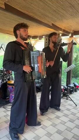 Two men stand performing music outdoors under a thatched roof structure. The man on the left is playing an accordion and singing. He has a beard and is wearing a black shirt and wide black pants. The man on the right is playing the violin and has a beard. He is wearing a black shirt and wide black pants. There are musical instruments and equipment visible in the background, including a drum set, speakers, and a music stand. The ground is tiled, and there are string lights overhead.