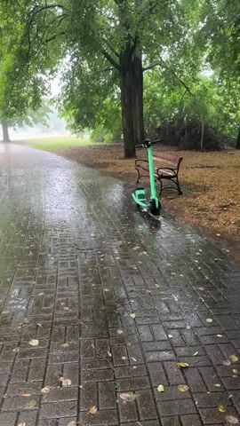This is a video of a rainy day in a park. The scene is a paved pathway winding through a lush green park with large trees. The pathway is wet and glistening from the rain, with fallen leaves scattered across it.

A bright green electric scooter is parked on the side of the pathway next to a wooden park bench. In the background, more trees and greenery are visible, with a slightly hazy atmosphere suggesting the rain. The overall mood is tranquil and somewhat melancholic due to the weather. The time of day appears to be daytime, but the overcast skies and rain obscure direct sunlight. There are no visible text elements in the video.