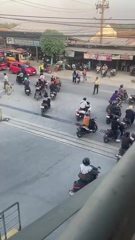 The video shows a street scene with a lot of motorcycle traffic. Several people are crossing the street, and some are waiting to cross. One person in a yellow vest appears to be directing traffic. There are also some cars and shops in the background. The scene appears to be in a busy urban area, possibly in Southeast Asia, given the prevalence of scooters and the style of buildings. The lighting suggests it is daytime, possibly late afternoon.
