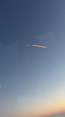 The video shows a bright orange streak across a blue sky. There is a small, bright circle to the left of the streak. The horizon is visible at the bottom of the frame with a gradient of soft colors. It appears to be a clear day, possibly at sunrise or sunset. The streak could be a contrail or a meteor. There are no people or objects identifiable in the video.