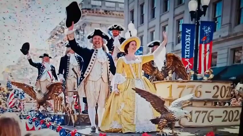 This is a video of a parade with confetti falling. In the foreground, a man and a woman in colonial-era attire are waving. The man wears a dark blue coat with white trim, a white wig, and a tricorn hat. The woman wears a large yellow gown with white ruffles and a matching hat. Behind them, several other men in similar colonial attire are also waving. The float they are on is decorated with eagles and banners that read "1776 - 2020". American flags are visible on the float and in the hands of people in the background. The environment appears to be an urban street with buildings lining the background.