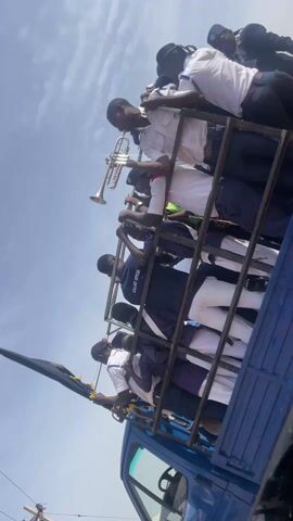 From inside a moving vehicle, we see a blue truck carrying people standing in its bed, which is enclosed by a metal cage. These people appear to be part of a parade band, with one man holding a trumpet and others wearing uniforms. The uniforms are a mix of white shirts, dark blue or purple trousers/skirts, and white sashes, and some bear the inscription "GALLANT OFFICER". A man is driving the truck, seen through the window. The sky is visible, with power lines present in the background. The surroundings suggest a developing area with partially constructed buildings. The video seems to have been taken during a procession or event.
