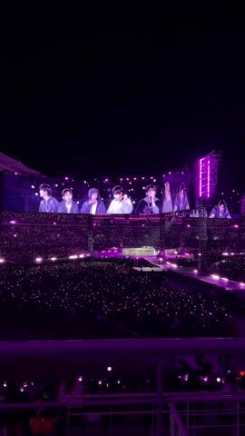 The video captures a BTS concert in a stadium at night. The audience is densely packed, holding up lights, creating a field of purple. On a large screen above the stage, the members of BTS are visible, appearing to be performing. The stadium is illuminated with purple lighting, enhancing the atmosphere of the event. The shot is taken from an elevated position, looking down towards the stage and audience. A portion of the stadium's infrastructure, including the railings and seats, is visible in the foreground.