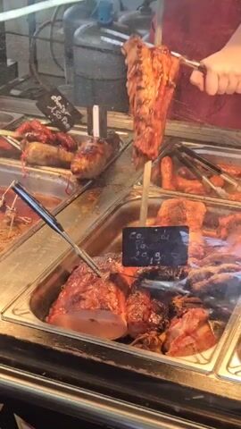 A close-up shot shows a metal display case filled with various cooked meats. A person wearing a red top is seen using tongs to manipulate a piece of grilled meat that's on a skewer. Other meats are also displayed in the case, including sausages and large cuts of pork, all in separate trays. Price tags are visible on each tray. Three gas cylinders are partially visible in the background. The environment is likely a deli or butcher shop.