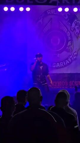 A musician is performing on stage in front of an audience. The musician is centered in the frame, wearing a dark outfit and a cap, holding a microphone and gesturing with their hands as they sing. Behind them is a backdrop featuring a logo with "THE RIDERS MC BULGARIA" written on it. The stage is lit with bright, colorful stage lights. The audience members are visible from the back, their silhouettes clear against the bright stage. The environment suggests a live music event or concert.