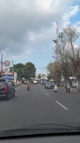 A street scene in what appears to be a tropical or subtropical climate. The view is from inside a vehicle, looking forward. The road is paved and has visible lane markings. Traffic consists of several vehicles, including cars, motorcycles, and scooters, moving in both directions. Along the sides of the road are buildings, some with signs, and trees. A tall communications tower is visible in the distance. The sky is partly cloudy.