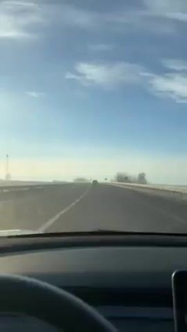 From the interior of a car, a person is driving on a straight highway with a car ahead of them. The sky is blue with streaky clouds. On the side of the road, there is snow and some bare trees. A small Dr. Marcus air freshener bottle is hanging from the rearview mirror. The car's dashboard shows the speed is 88 and there is 13km to the destination.