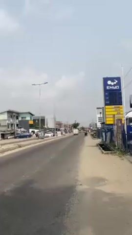 A long, straight paved road with some cracks is shown in a bright, daytime, outdoor shot. The sky is slightly overcast. On the right side of the road is an Enyo gas station sign. The price displays are all set to 888.88. A dirt shoulder runs alongside the paved road. In the background are various low-rise buildings and trees. Several vehicles pass along the road including cars, motorcycles and a yellow tricycle (tuk-tuk). A motorcycle sits in the lower left corner of the frame. Overhead are streetlights.