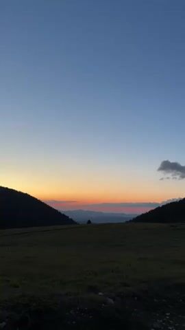 The video shows a time-lapse of a sunset over a mountainous landscape. The sky transitions from a deep blue at the top to a band of orange and pink near the horizon. Dark silhouettes of hills and mountains frame the scene, with a grassy meadow in the foreground. A few scattered clouds drift across the sky. There are no people or man-made objects visible. The overall mood is serene and natural.