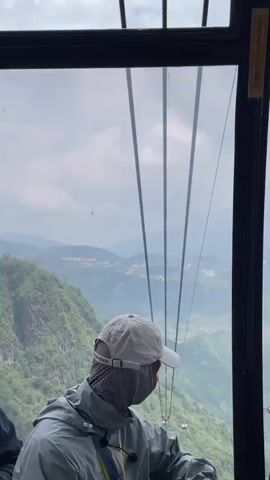 The video shows the interior of a cable car ascending a mountain. A person wearing a light-colored baseball cap and a protective face covering is looking out the window at a green, mountainous landscape. Several cables are visible stretching across the frame. Another person is visible in the foreground, looking at their phone. The cable car has a red interior trim and a window that is partially open. The sky is cloudy.