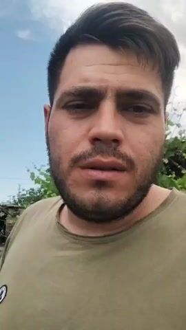 A medium close-up shot of a man with dark hair and beard looks directly at the camera. He is wearing a greenish-brown t-shirt. He is outdoors, with a blue sky and some trees in the background. The man speaks and smiles at the camera.