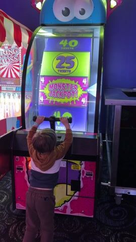A small child is seen from the back, playing a game at an arcade. The child wears a brown and white striped hoodie and brown pants. The arcade game has a large, blue-colored topper with cartoonish eyes. Below the topper is a tall rectangular screen which displays digital images of tickets and the words "MONSTER JACKPOT!" The game console is decorated with a pink, purple, and yellow cartoon monster theme. The child stands on their toes and pulls a t-shaped handle back and forth, engaging in the arcade game. The background features typical arcade aesthetics: other arcade games with bright colorful decorations and lighting.