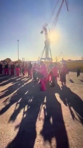 The video depicts a group of people, dressed in pink suits, playing musical instruments outside. The background features industrial equipment, including large cranes, and the scene is bright with sunlight. The camera moves slightly, showing the group from various angles as they play. The overall atmosphere appears festive or celebratory.