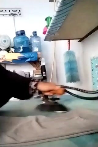The video shows a person ironing a light-colored piece of fabric, likely clothing, on an ironing board. The person is holding an iron with a black handle and a black cord that extends out of frame. The ironing board is set up in front of a wall with shelves holding various items, including water bottles, a fan, and cleaning supplies. A blue duster hangs from the underside of the ironing board. The lighting appears to be artificial and relatively dim.