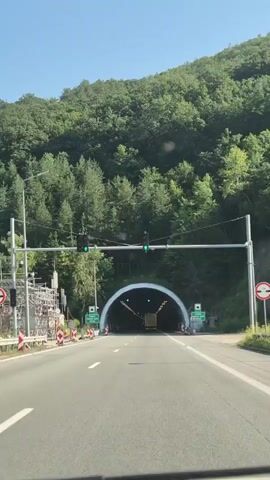 A vehicle approaches a tunnel entrance on a sunny day. The tunnel is wide and arched, illuminated by lights. Green traffic signals are visible above the road, indicating the tunnel is open. The surrounding landscape is mountainous and covered in dense green trees. The road leading to the tunnel has multiple lanes and is marked with white lines. Construction barriers and scaffolding are present on the left side of the road near the tunnel entrance. The video is filmed from inside a vehicle, likely the driver's perspective.