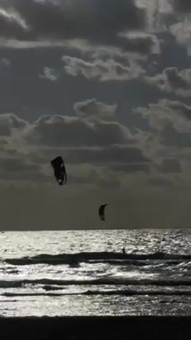 The video shows a silhouette of two kitesurfers riding in the ocean on a cloudy day. The sky is filled with dark, textured clouds, and the sun reflects brightly off the surface of the water. The kitesurfers are small figures in the distance, each attached to a kite that is visible against the sky. The waves are small and choppy. The beach in the foreground is dark and undefined, adding to the overall silhouette effect.