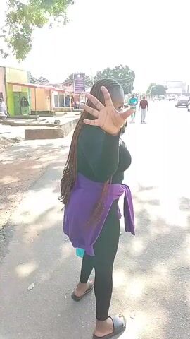 The video depicts a woman walking away from the camera on a paved road. She is wearing a black long-sleeved shirt, black leggings, a short purple skirt, and dark sandals. She has long dark hair styled in braids. A red purse is slung over her shoulder, and she is carrying a blue shopping bag with a graphic on it. The woman initially holds up her hand as if to block the camera's view, then proceeds to walk away. In the background, other people are walking in the opposite direction, and there are parked cars and buildings along the road. The scene appears to be a sunny day in an urban or suburban environment.