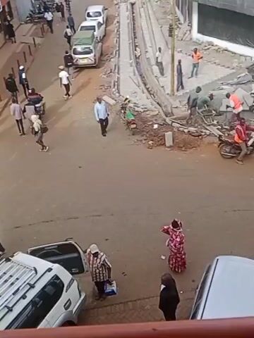 Here is a brief description of the video:

The video shows a street scene. The camera is angled down, providing an overhead view. There are several people walking and standing on the street, along with parked cars and motorcycles.  A yellow bus with the name "St. Peter's High Class Academy Kapsabet" printed on its side pulls into the scene and stops.  Construction work is visible on one side of the street.