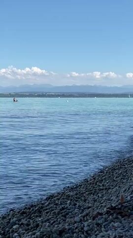 A wide shot captures a tranquil body of water under a bright blue sky dotted with white clouds. In the distance, hazy mountains form a picturesque backdrop. The foreground is dominated by a pebble beach, with gentle waves lapping at its edge. A lone kayaker is visible in the middle ground, paddling across the clear blue water. The scene conveys a sense of peacefulness and natural beauty.