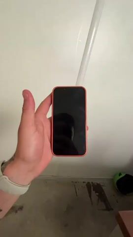 A hand holds a bright orange smartphone vertically in front of a white wall and a grey concrete floor. The phone's screen is black, reflecting the person's face and the ceiling light. The hand turns the phone to reveal its back, which has a distinct camera module with two lenses arranged diagonally within a raised, darker orange rectangular section at the top. The Apple logo is visible in the center of the lower part of the phone's back. The person is wearing a white fitness tracker on their left wrist, and a tattoo of barbed wire is visible on their forearm. In the background, a white pipe runs diagonally along the wall, and a glimpse of a green object, possibly a helmet, is seen on the floor. The video shows the phone being rotated to display both its front and back.