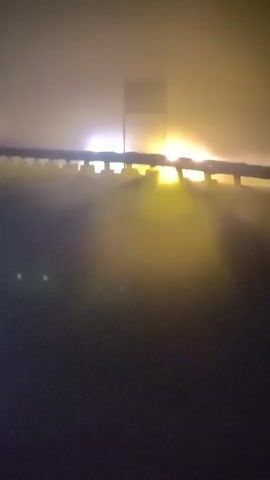 A vehicle approaches and passes over a bridge with fog obscuring the view. The vehicle's headlights illuminate the fog as it moves. There is a railing on the side of the bridge and a large signpost in the distance.