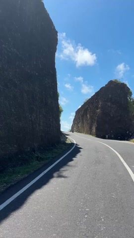 The video shows a straight, paved road running between two tall, dark rock formations. A person on a scooter drives down the road towards the viewer. The sky is blue with scattered white clouds.