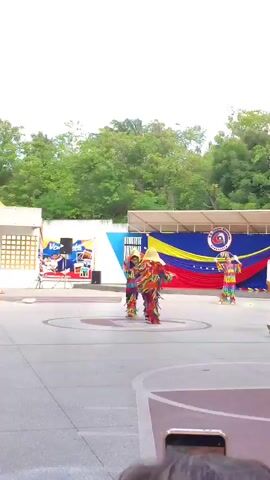 The video shows a group of dancers in colorful costumes performing in an outdoor space. They are dressed in full-body outfits with streamers and masks, reminiscent of traditional festival costumes.  The performance appears to be taking place on a basketball court, judging by the painted lines on the ground. In the background, there's a Venezuelan flag with a logo, suggesting this is a cultural event. Behind the dancers, there is a building with brick accents and a person observing from a balcony. The day is bright but overcast.