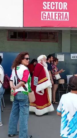 The video shows a man dressed as Santa Claus standing near a group of people at a polling station. The polling station is labeled "Sector Galería" and "Mesa 493". Several individuals are seen interacting at tables, seemingly engaged in voting-related activities. One woman is wearing a shirt with the colors of the Palestinian flag. The area appears to be an outdoor covered space.