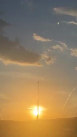 The video shows a sunset, with the sun low on the horizon, casting a golden glow over the scene. The sky is partly cloudy. A white car is briefly visible passing by on a road. Power lines and light poles are also visible in the distance. A jet stream is visible.