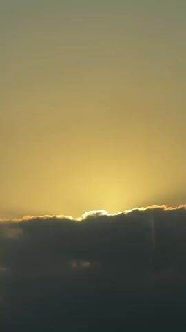 The video shows a view from inside a bus, looking out a window. In the foreground, a black object, possibly a camera lens cap, rests on the seat. The background shows a landscape with mountains and fields under a sky with clouds and a setting sun. The sun's rays are peeking through the clouds, creating a dramatic effect. The video also includes close-ups of the clouds and the sun.