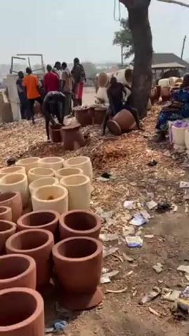 The video shows a marketplace or workshop environment where wooden mortars and pestles are being crafted or sold. Several men are visible, some working on the mortars, with one man sitting on a mortar, possibly smoothing or shaping it. The ground is covered in wood shavings and debris. There are stacks of finished and unfinished mortars in various sizes and colors. Other people are walking around, seemingly browsing or engaged in transactions. The overall impression is one of a busy, industrious setting focused on the production of traditional wooden tools.