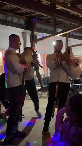 Under a wooden-beamed structure with colored lights, three men stand playing bagpipes, the two at the front wearing white shirts with decorative embroidery at the cuffs. They are playing for an audience, a few people visible at the edges of the frame, in a room set up with tables and chairs. The floor is tiled. The musicians are dressed in dark pants and sneakers. The bagpipes are light in color with long wooden pipes.