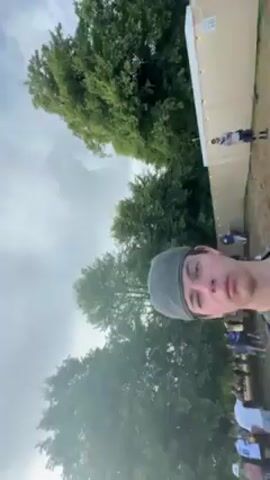 Here is a concise description of the video:

The video shows a person filming themselves at a music festival. The camera pans around to show a large grassy field with trees, people walking around, and colorful flags and decorations. It appears to be a sunny day with some clouds in the sky.