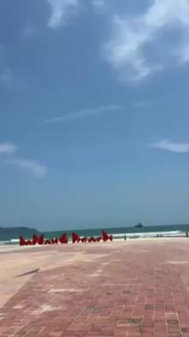The video shows an outdoor scene, likely a beachside event space. A wide paved area, patterned with red bricks, stretches towards the ocean. In the distance, the ocean is visible with small waves breaking along the shoreline under a bright blue sky dotted with a few wispy clouds. A small island or headland is seen in the background.

On the paved area, a series of red, angular objects stand in a row. They appear to be large, decorative elements forming a word or a visual pattern. To the left of these objects, a large stage has been erected. The stage has a white roof supported by a metal frame, and it contains sound equipment and banners. The atmosphere suggests preparations are underway for an event or performance. The scene is brightly lit, indicating a sunny day.