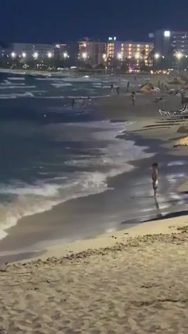 The video captures a nighttime scene at a beach, likely in Cala Millor, Spain, given the context. The beach is illuminated by the lights of buildings in the background, suggesting a bustling resort area. The sea is active with moderate waves breaking on the shore, creating a dynamic contrast with the calm, sandy foreground.

The primary subject is a young child standing in the shallow water, appearing to be enjoying the evening at the beach. The child is wearing light-colored shorts and is facing away from the camera, looking out at the sea. Other people are visible in the distance, scattered along the beach and in the water, engaged in various activities. Some are walking, while others appear to be relaxing on loungers under thatched umbrellas. The presence of these umbrellas and loungers indicates a developed tourist destination.

The time of day is clearly night, as evidenced by the dark sky and the artificial lights from the buildings and potentially streetlights. The weather appears calm, with no indication of rain or strong winds, allowing for beach activities to continue into the evening. The sand in the foreground is detailed, with shadows cast by unseen objects, possibly from the thatched umbrellas. The overall mood is peaceful and relaxed, typical of a pleasant evening at a seaside resort. There is no visible text in the video.