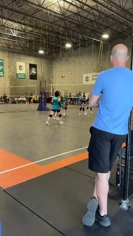 The video shows a man watching a volleyball game. He is standing to the side of the court with his arms crossed, wearing a light blue t-shirt and black shorts. The game is between two teams of young girls wearing teal and black uniforms. The setting is an indoor gymnasium with high ceilings and visible metal support beams. There are spectators seated in the background, and another person with blonde hair walks by the camera. The man appears to be focused on the game in progress.