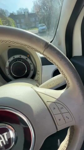 A woman in a white car, wearing sunglasses and a gray sweater, rests her head against the car door frame, looking up. The interior of the car has red seats and a white steering wheel. The car is parked, and a tree with white blossoms is visible against a blue sky. The camera angle shifts slightly, focusing more on the tree and then back to the woman in the car.