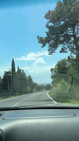 The video is a first-person perspective from inside a car, looking out through the windshield. The car is driving on a paved road that curves to the right. On either side of the road, there are trees and green foliage. The sky is blue with scattered white clouds. In the distance, the road continues, and there are a few other cars visible. The interior of the car, including the dashboard, is visible at the bottom of the frame. The overall scene suggests a sunny day driving through a rural or suburban area with a lot of greenery.