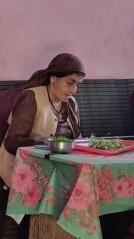 An elderly woman with a headscarf is sitting at a table covered with a floral tablecloth. She is wearing a light brown vest over a brown patterned shirt. In front of her on the table are a metal bowl filled with chopped green herbs and a red cutting board with more green herbs. Her right hand is reaching towards the herbs on the cutting board. The background shows a pink wall and a couch with striped cushions.