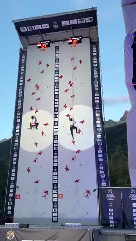 The video shows a competitive rock climbing event taking place outdoors during daylight. Two athletes are scaling a tall, white artificial climbing wall. The wall is divided into two identical vertical lanes, each marked with numerical height indicators and a large letter "A" and "B" at the bottom. Red climbing holds are strategically placed on the wall. Above the climbing lanes, digital timers are visible, displaying the athletes' times. Both climbers are equipped with harnesses and ropes for safety, and appear to be in mid-climb. The background reveals a mountainous landscape under a clear blue sky, suggesting the competition is held in a scenic, high-altitude location. Logos associated with "WORLD CLIMBING" and "THE NORTH FACE" are displayed on banners and the top structure of the climbing wall.