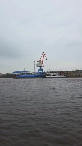 The video presents a panoramic view of a river and its adjacent industrial bank on an overcast day. The camera slowly pans from left to right, revealing various elements of the scene.

In the initial frames, a prominent industrial port crane, featuring an orange boom and a blue base, stands at a dock on the left side of the riverbank. Moored below the crane is a long, flat-bottomed vessel or barge covered with a blue tarp. Beside it, a white, multi-decked passenger boat or ferry with red accents is also docked. Behind these structures, several light-colored buildings with blue roofs are visible, suggesting port facilities.

The river, occupying the lower half of the frame, is dark and appears to have a choppy surface, indicating a current. The sky above is uniformly gray and overcast, suggesting a cloudy day with soft, diffused light.

As the camera pans right, the crane and boats gradually move out of view. The distant riverbank reveals a long, dark, textured retaining wall or embankment. Beyond this, a strip of green land is visible, with what appears to be a road or railway line running parallel to the river. Faintly visible on this distant strip are what could be trucks or cargo containers, along with a tall, slender metal tower. No discernible people are visible, and there is no clear text on any of the objects or buildings. The overall setting is that of an active industrial river port.