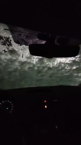 From the interior of a vehicle at night, heavy snow falls onto the windshield. The wipers briefly swipe away the snow, revealing a dimly lit exterior. The dashboard is dark, with a few illuminated indicators. A hand is briefly visible on the steering wheel. The rearview mirror is prominent in the center of the frame. The overall environment appears to be quiet and snowy.