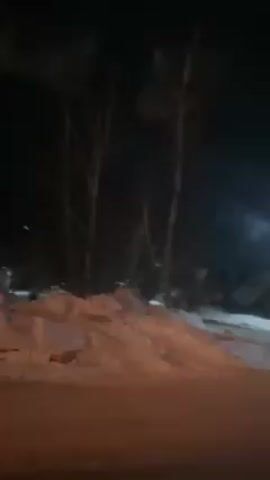 The video depicts a snow-covered street at night. There are trees with bare branches visible against the dark sky. Snow is piled high along the edge of the road. Streetlights illuminate the scene, casting shadows and highlighting the textures of the snow. A person is walking on the sidewalk in the background. The camera appears to be inside a vehicle.