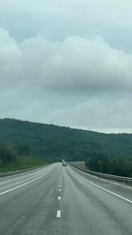 The video shows a view from a car driving on an asphalt road. The road has white dashed lines marking the lanes. On the left side of the road, there is a grassy shoulder with trees and a dense forest covering a hill. On the right side, there is a guardrail and more trees. In the distance, two trucks are visible on the road. The sky is overcast with gray clouds. The overall scene depicts a journey through a natural, forested landscape.