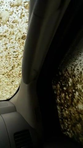 The video shows the interior of a vehicle, looking out through heavily splattered windows. The side window and what appears to be the sunroof or upper part of the windshield are covered in thick, dark brown mud and dirt. The mud dramatically obscures the view outside. The sound of a running engine is audible throughout the clip, suggesting the vehicle is currently driving or has just driven through a very muddy area. The ambient light indicates it is daytime, with a bright, possibly overcast, sky visible through the mud. The location context provided is Manavi, Georgia.