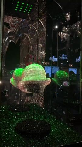 A crystal-encrusted bowler hat is displayed in a glass case. The hat is illuminated by green lights, and the case is surrounded by mirrors, creating a kaleidoscope effect. The hat sits on a pedestal in front of a background of mirrored surfaces with ornate, crystal-covered archways. The bottom of the display case is covered in black material and green glitter. The video is a close-up shot of the hat from a slightly low angle, with no people visible in the frame.