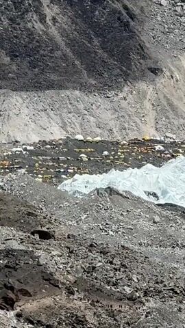 The video presents a striking aerial view of a bustling mountain base camp, likely Everest Base Camp, set against the backdrop of towering Himalayan peaks.

Initially, the camera focuses on a dense cluster of colorful tents—white, yellow, orange, and blue—spread across a rocky, uneven terrain. This tent city is situated directly beside a prominent expanse of white glacier ice, with steep, barren mountain slopes rising immediately behind it. Small, dark figures, identifiable as people, can be seen traversing the rocky paths in the foreground, indicating active trekking or movement around the camp.

As the camera slowly zooms out, the vastness of the surrounding landscape is revealed. The base camp and glacier become smaller within the frame, emphasizing the immense scale of the rocky, gray and brown mountains that stretch into the distance. The final frames offer a panoramic vista, showcasing a majestic, snow-capped mountain peak dominating the horizon under a clear, bright blue sky with a few wispy white clouds. The foreground and mid-ground are characterized by rugged, rocky terrain and visible glacier sections.

The scene depicts a clear, sunny day, highlighting the harsh yet beautiful high-altitude environment. The abundance of tents and the dramatic mountainous setting are strong indicators of a major mountaineering hub in the Khumbu region of Nepal.