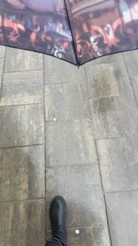 The video shows a person standing under an umbrella on a wet, paved walkway. The umbrella has a decorative image of a canal scene, possibly in Amsterdam. The person is wearing a black boot. The ground is wet from rain, and the walkway is made of gray rectangular pavers. The view is from the person's perspective, looking down at the ground and out past the edge of the umbrella. Beyond the walkway is a strip of grass and a road. The overall impression is of a rainy day.